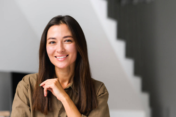 Portrait of charming elegant asian woman with hand under chin smiling at the camera, sitting in the modern house, wearing domestic clothes - Foto, afbeelding