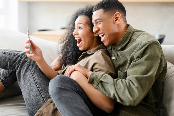 Excited and overjoyed multiracial young couple sitting on the couch at home and holds smartphone. Man and woman celebrating good news or win - Fotó, kép