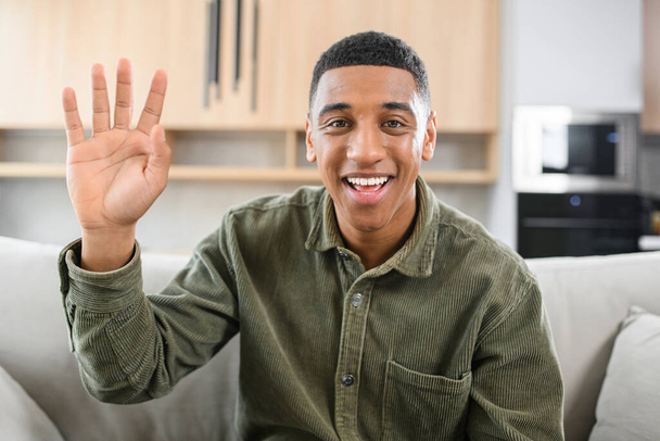 Portrait of happy young multiracial guy sitting at desk and waving at camera. Smiling man in casual shirt involved virtual meeting - Photo, Image