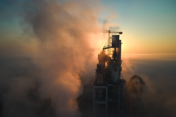 Αεροφωτογραφία του εργοστασίου τσιμέντου με την υψηλή δομή του εργοστασίου τσιμέντου και τον πυργογερανό στη βιομηχανική μονάδα παραγωγής το ομιχλώδες βράδυ. Παραγωγή και έννοια της παγκόσμιας βιομηχανίας - Φωτογραφία, εικόνα