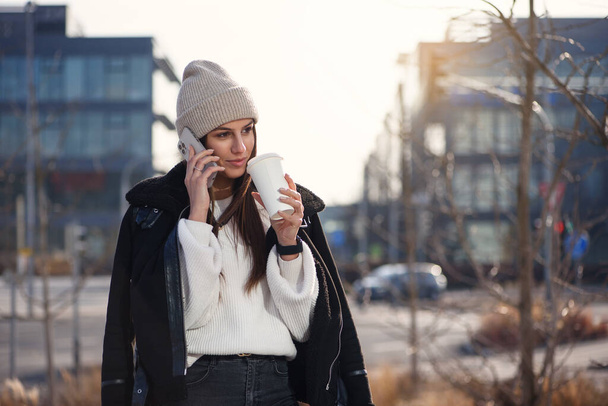 Femme souriante avec téléphone intelligent et café pour aller debout à l'extérieur et parler. Concept de style de vie moderne. - Photo, image