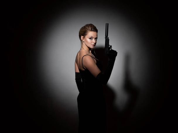 dangerous spy. A femme fatale in a black dress with an open back holds a pistol - Foto, afbeelding