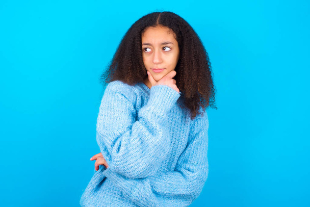 Задумчивая улыбающаяся афроамериканская девочка-подросток с афро-прической в синем свитере на синем фоне держит руку под подбородком, смотрит прямо в камеру, слушает что-то интересное. Концепция молодежи. - Фото, изображение