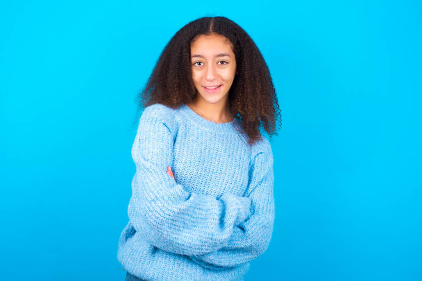 Афроамериканская девочка-подросток с афро-прической в синем свитере на синем фоне, счастливая улыбаясь и скрещивая руки, уверенно смотрящая в камеру. Позитивный и уверенный человек. - Фото, изображение