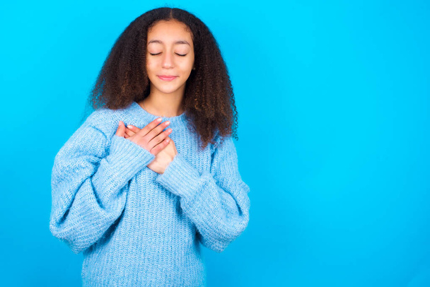 African American teenager girl with afro hair style wearing blue sweater over blue background closes eyes and keeps hands on chest near heart, expresses sincere emotions, being kind hearted and honest. Body language and real feelings concept. - Photo, Image