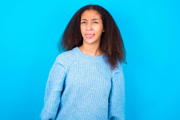 Langage corporel. Dégoûté stressé fille adolescente afro-américaine avec une coiffure afro portant un pull bleu sur fond bleu, le visage froncant des sourcils, démontrant une aversion pour quelque chose. - Photo, image