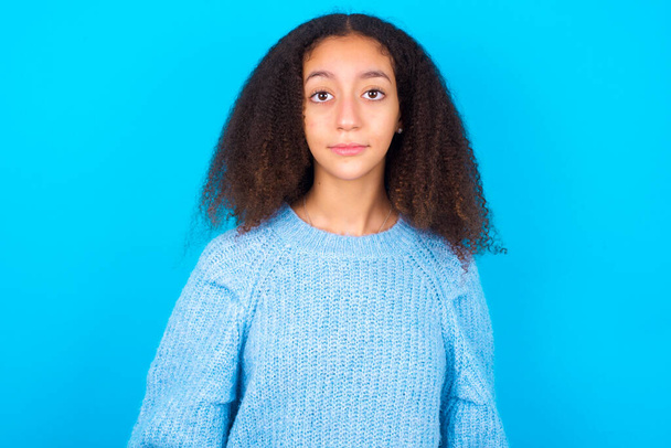 Iloinen afroamerikkalainen teini-ikäinen tyttö, jolla on afro-kampaus yllään sininen pusero sinisellä taustalla, katsoo kameraan ja ajattelee jotain. Molemmat kädet alas, neutraali ilme. - Valokuva, kuva