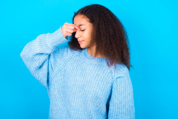 Very upset, African American teenager girl with afro hair style wearing blue sweater over blue background touching nose between closed eyes, wants to cry, having stressful relationship or having troubles with work - Foto, imagen