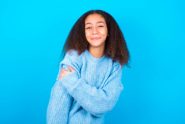 People, lifestyle, youth and happiness concept. Shy pretty African American teenager girl with afro hair style wearing blue sweater over blue background, feeling happy hugging herself. - Φωτογραφία, εικόνα
