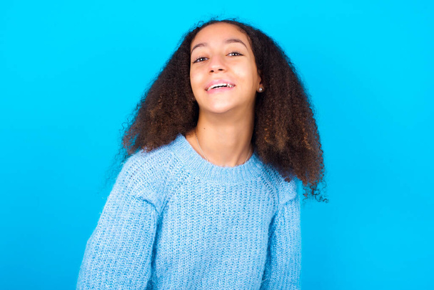 African American έφηβος κορίτσι με αφρο στυλ μαλλιών φορώντας μπλε πουλόβερ πάνω από το μπλε φόντο με ευρύ χαμόγελο, δείχνει λευκά δόντια, αίσθημα χαράς έχοντας ρεπό. - Φωτογραφία, εικόνα