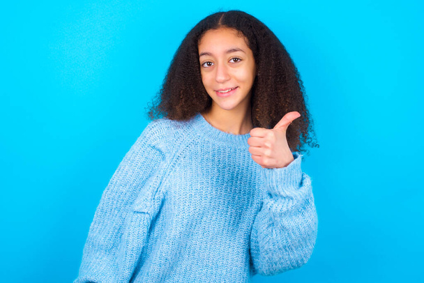 Afrikai amerikai tinédzser lány afro frizura visel kék pulóver felett kék háttér így hüvelykujj fel gesztus, jó Job! Pozitív emberi érzelem arckifejezés testbeszéd. - Fotó, kép