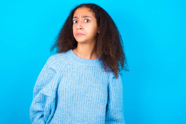 African American teenager girl with afro hair style wearing blue sweater over blue background with snobbish expression curving lips and raising eyebrows, looking with doubtful and skeptical expression, suspect and doubt. - Photo, Image
