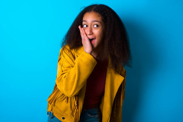 Afrikkalainen amerikkalainen teini tyttö afro hiukset tyyli yllään keltainen takki yli sininen tausta innoissaan katsellen sivulle käsin kasvot. Mainonnan ja hämmästyksen käsite. - Valokuva, kuva