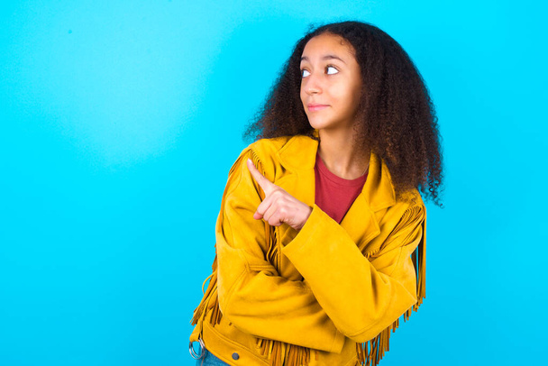 Ritratto di adolescente afro-americana con acconciatura afro che indossa giacca gialla su sfondo blu in posa sulla macchina fotografica con look complicato, presentando il prodotto con indice. Concetto di pubblicità. - Foto, immagini