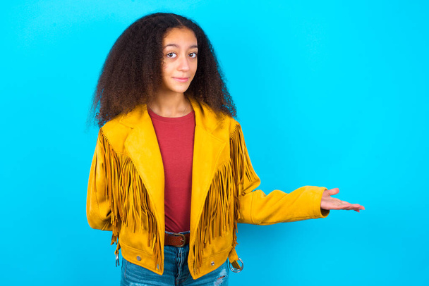 アフリカ系アメリカ人のティーンの女の子の肖像とともにアフロヘアスタイル身に着けています黄色のジャケット上に青の背景とともにアームアウトで歓迎ジェスチャー. - 写真・画像