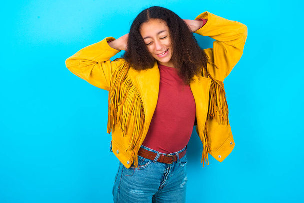 Afrikkalainen amerikkalainen teini tyttö afro hiukset tyyli yllään keltainen takki yli sininen tausta rentouttava ja venyttely, kädet pään ja kaulan takana hymyillen onnellinen - Valokuva, kuva