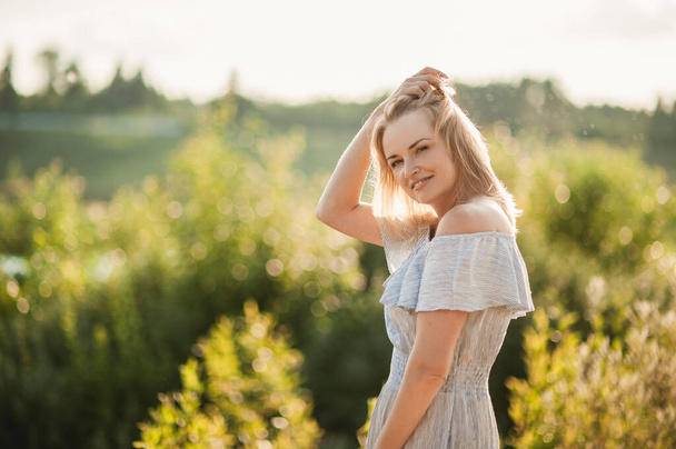Portrait of a beautiful blonde woman in a summer dress at sunset among the beautiful nature. The girl adjusts her hair and looks into the camera. Space for text. - Foto, immagini