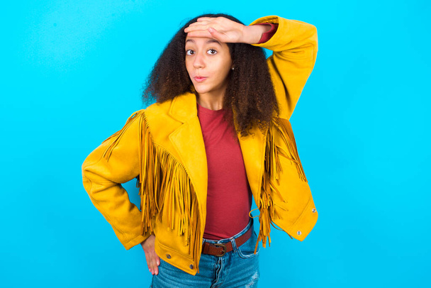 Африканська дівчинка-підліток з афроамериканським волоссям, одягнена в жовтий піджак над синім фоном, витирає чоло руками, роблячи жести феєм, виражає полегшення, відчуваючи себе щасливим, що він запобіг величезним катастрофам. Вона була досить близько. - Фото, зображення