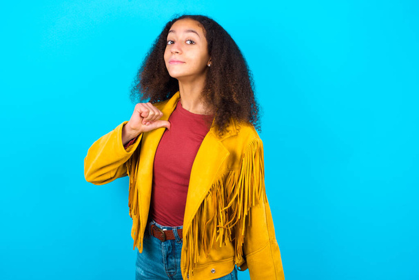 Primer plano de la alegre adolescente afroamericana con estilo afro pelo usando chaqueta amarilla sobre fondo azul se ve alegre, satisfecho y confiado, apunta a sí mismo con el pulgar. - Foto, Imagen