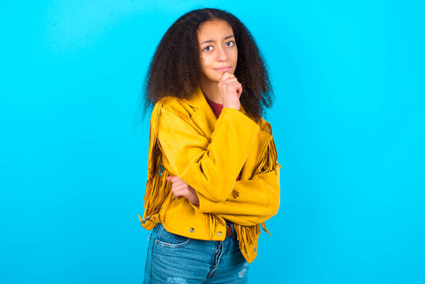 Afro-amerikanisches Teenager-Mädchen mit Afro-Frisur trägt gelbe Jacke über blauem Hintergrund mit der Hand unterm Kinn und schaut mit zweifelndem und skeptischem Gesichtsausdruck seitwärts, misstrauisch und zweifelnd. - Foto, Bild