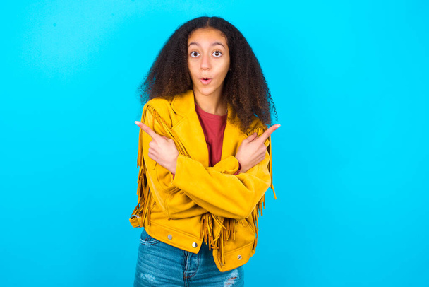 青い背景の上に黄色のジャケットを着てアフロの髪のスタイルを持つ混乱アフリカ系アメリカ人の10代の少女は、 2つの方法の間を選択し、交差した手で両側のポイントは、疑問を感じている。君の助言が必要だ. - 写真・画像