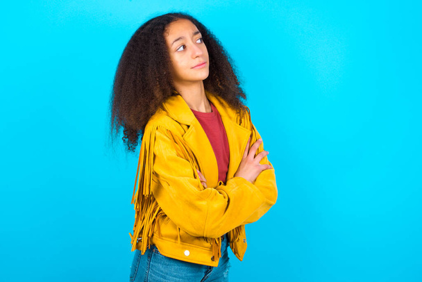 Afbeelding van overstuur Afro-Amerikaanse tiener meisje met afro haar stijl dragen gele jas over blauwe achtergrond met gekruiste armen. Kijkend met teleurgestelde uitdrukking terzijde na het luisteren naar slecht nieuws. - Foto, afbeelding