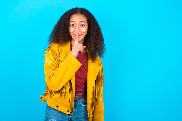 Mosolygó afro-amerikai tinédzser lány afro haj stílus visel sárga kabát a kék háttér teszi csöndes gesztus, tartja előujját az ajkak felett elrejti a titkot. Hallgasson, kérem.. - Fotó, kép