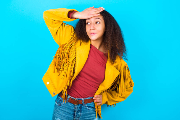 青い背景に触れる額の上に黄色のジャケットを着てアフロヘアスタイルを持つアフリカ系アメリカ人の10代の少女は、何か驚くべきことを聞くと、うれしい良いニュースを受け取る、安心感。トラブルに巻き込まれそうになった. - 写真・画像