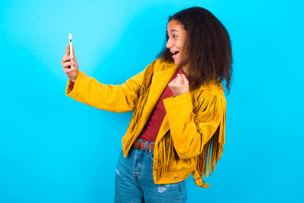 Fröhliches, fröhliches afroamerikanisches Teenager-Mädchen mit Afro-Frisur in gelber Jacke vor blauem Hintergrund, das per E-Mail gute Nachrichten erhält und im Stehen und beim Anblick des Mobiltelefons Erfolge feiert. - Foto, Bild