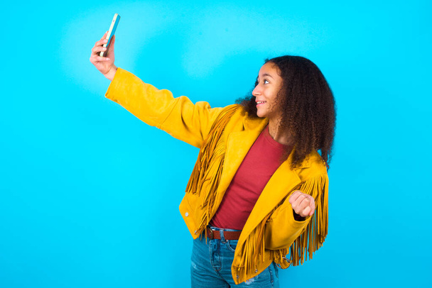 Afro saçlı, mavi arka planda sarı ceketli bir kızın portresi. Arkadaşlarına ve takipçilerine göndermek için selfie çekiyor ya da sosyal medyasına yüklüyor.. - Fotoğraf, Görsel