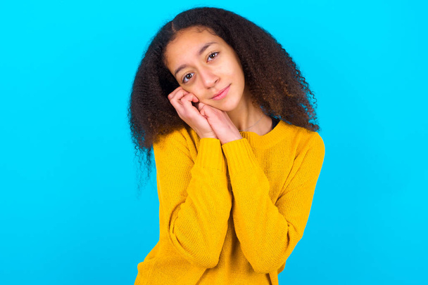 Charming serious African American teenager girl with afro hair style wearing yellow sweater over blue background  keeps hands near face smiles tenderly at camera - Photo, Image