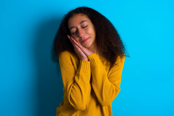 African American teenager girl with afro hair style wearing yellow sweater over blue background leans on pressed palms closes eyes and has pleasant smile dreams about something - Foto, imagen