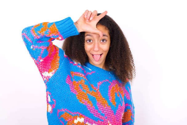 afro americano adolescente ragazza con afro capelli stile indossando vintage colorato maglione su bianco sfondo gesti con dito sulla fronte rende perdente gesto rende divertente di persone mostra lingua - Foto, immagini
