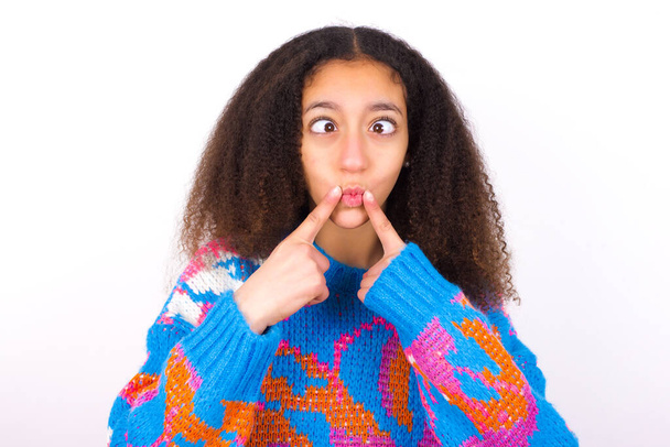 Chica adolescente afroamericana con el pelo rizado en estilo afro usando suéter colorido vintage sobre fondo blanco cruza los ojos y hace que los labios de pescado mueca divertida - Foto, imagen