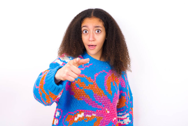 Shocked African American teenager girl with afro hair style wearing vintage colourful sweater over white background points at you with stunned expression - Photo, Image