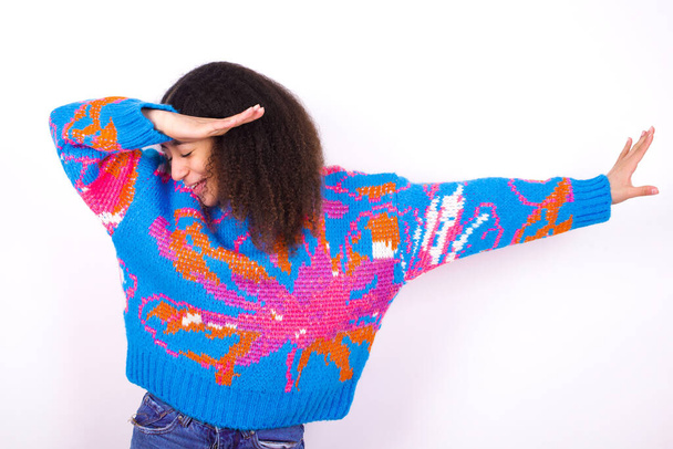 Foto de chica adolescente afroamericana funky con el pelo rizado en estilo afro usando suéter colorido vintage sobre fondo blanco mostrar disco mover dab - Foto, Imagen
