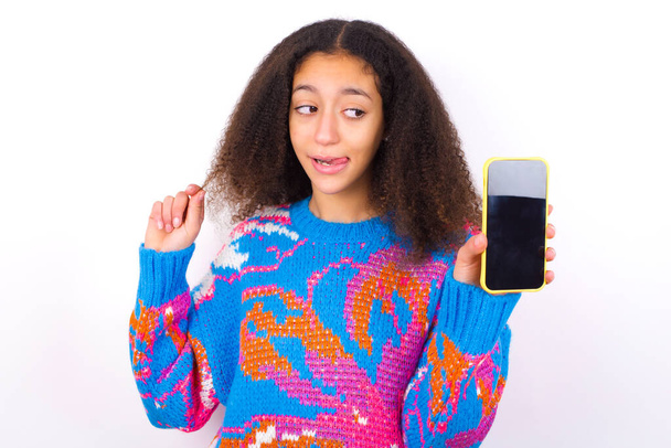 Photo of nice pretty African American teenager girl with curly hair in afro style wearing vintage colorful sweater over white background demonstrate phone screen hold hair tails - Foto, Bild