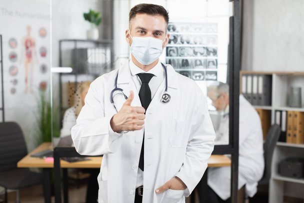 Doctor in face mask and lab coat posing at workplace - Foto, afbeelding