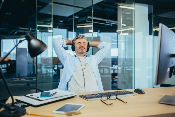 Senior and experienced business man resting at work during a break listening to music from headphones, manager working in a modern office - Photo, image