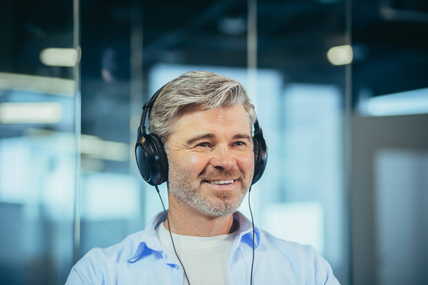 Close-up portrait of a man with gray hair listening to music in big headphones, a man working in the office - Photo, image