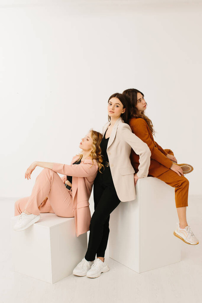 Model kadınlar, modaya uygun pastel ceketler ve şık spor ayakkabılar giyip stüdyoda beyaz bir arka planda küp üzerinde poz veriyorlar.. - Fotoğraf, Görsel