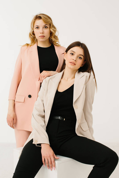 Model-Frauen in modischen Pastellblazern und Hosen mit stylischen Turnschuhen posieren auf einem Würfel auf weißem Hintergrund im Studio. - Foto, Bild