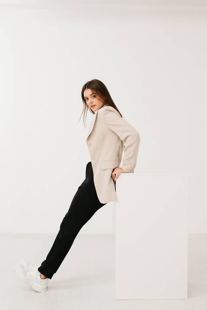Jonge vrouw draagt trendy pastel blazer en broek met stijlvolle sneakers zittend op kubus tegen witte achtergrond in studio. - Foto, afbeelding
