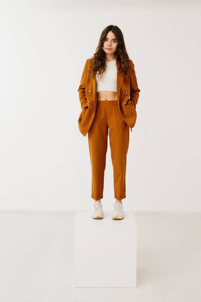 Young woman dressed in fashionable pastel blazer and pants with stylish sneakers standing on a cube on a white background in the studio. - Photo, image