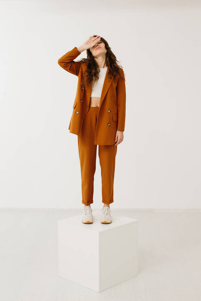Молодая женщина одета в модный пастельный пиджак и брюки со стильными кроссовками, стоящими на кубе на белом фоне в студии. - Фото, изображение