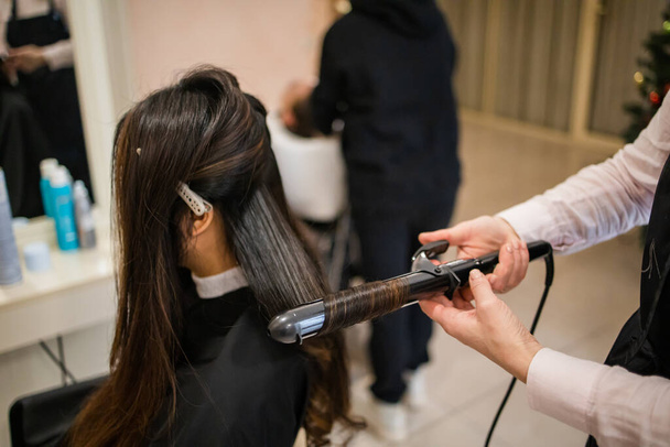 Hairdresser close-up makes a hairstyle with curling iron. - Photo, Image