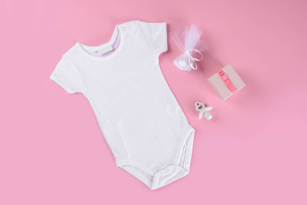 Beyaz pamuklu bebek tulumu, pembe renkli arka planda. Mesaj veya logo için boş bir yer. Bebek kıyafetleri, seramik meme ucu ya da bebek mankeni, hediye kutusu ve konfetiyle düzleştirilmiş. - Fotoğraf, Görsel