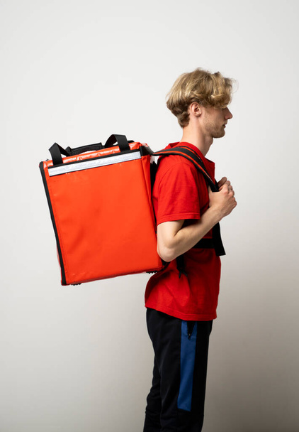 Fast Food Delivery Service Concept. Side view of smiling male courier wearing red uniform and holding thermal backpack bag isolated on studio background. - Photo, Image
