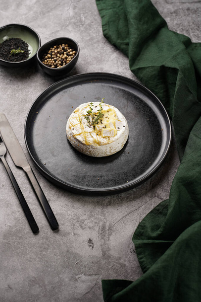 Oven baked camembert cheese with black pepper, sesame seeds on black plate, grey concrete surface. Homemade grilled brie with thyme, dark table setting - Фото, изображение