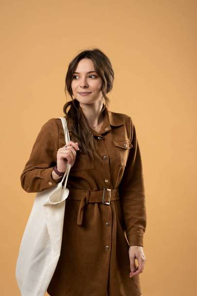 Happy smiling young woman in brown dress holding reusable string bag with groceries over orange background. Zero waste, plastic free concept. Eco lifestyle. Eco shopping. - Фото, зображення